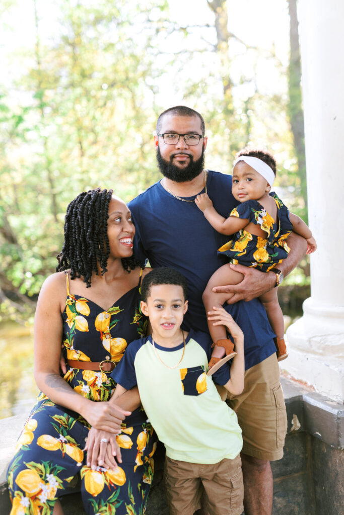 Family of four smiles at each other in matching lemon outfits during family session at gazebo over Lake Clara Meer in Atlanta, Georgia