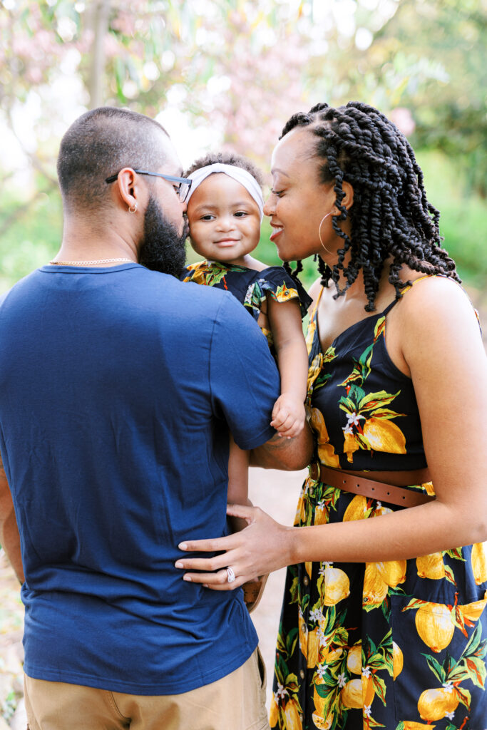 Mom and dad kiss their baby girl in the shade next to lake at Piedmont Park