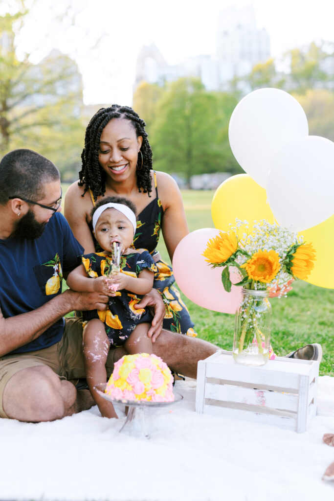 Family encourages baby girl to eat first birthday smash cake during milestones photo session