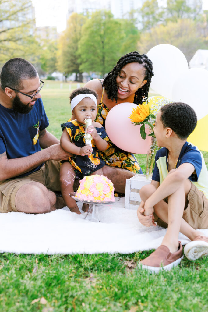 Family encourages baby girl to eat first birthday smash cake during milestones photo session at Piedmont Park in Atlanta, Georgia