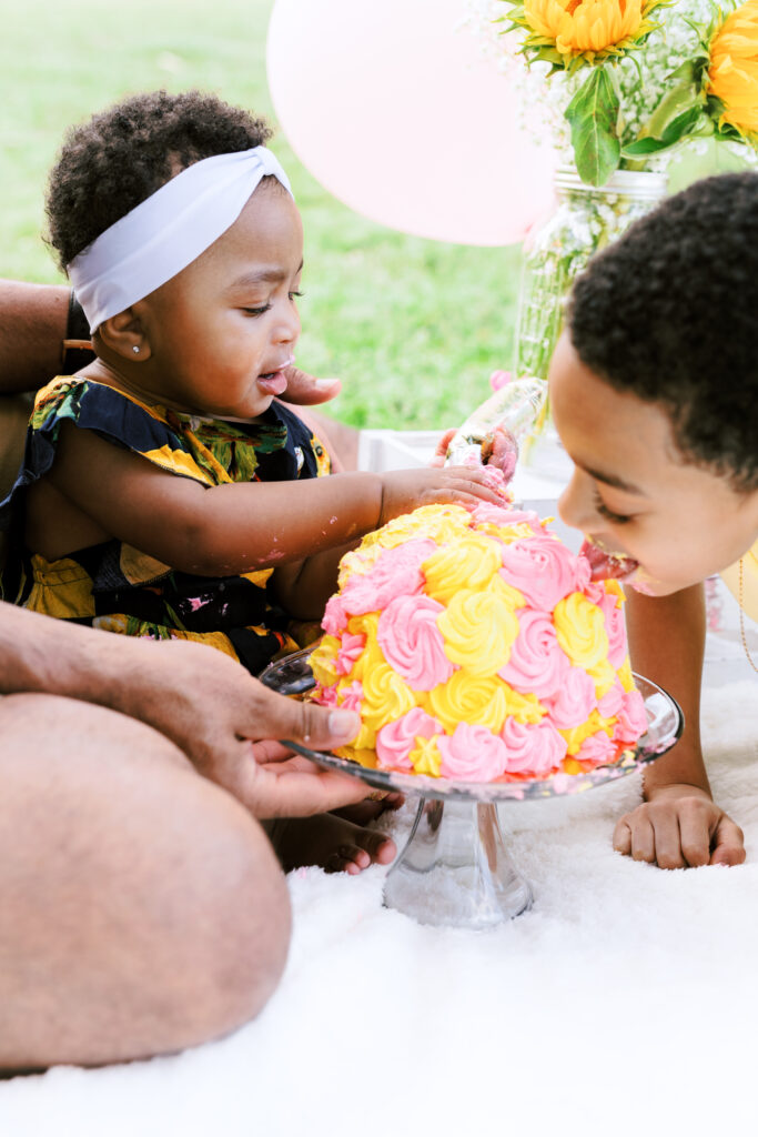 Baby smashes first birthday smash cake and big brother licks icing during milestones photo session at Piedmont Park in Atlanta, Georgia