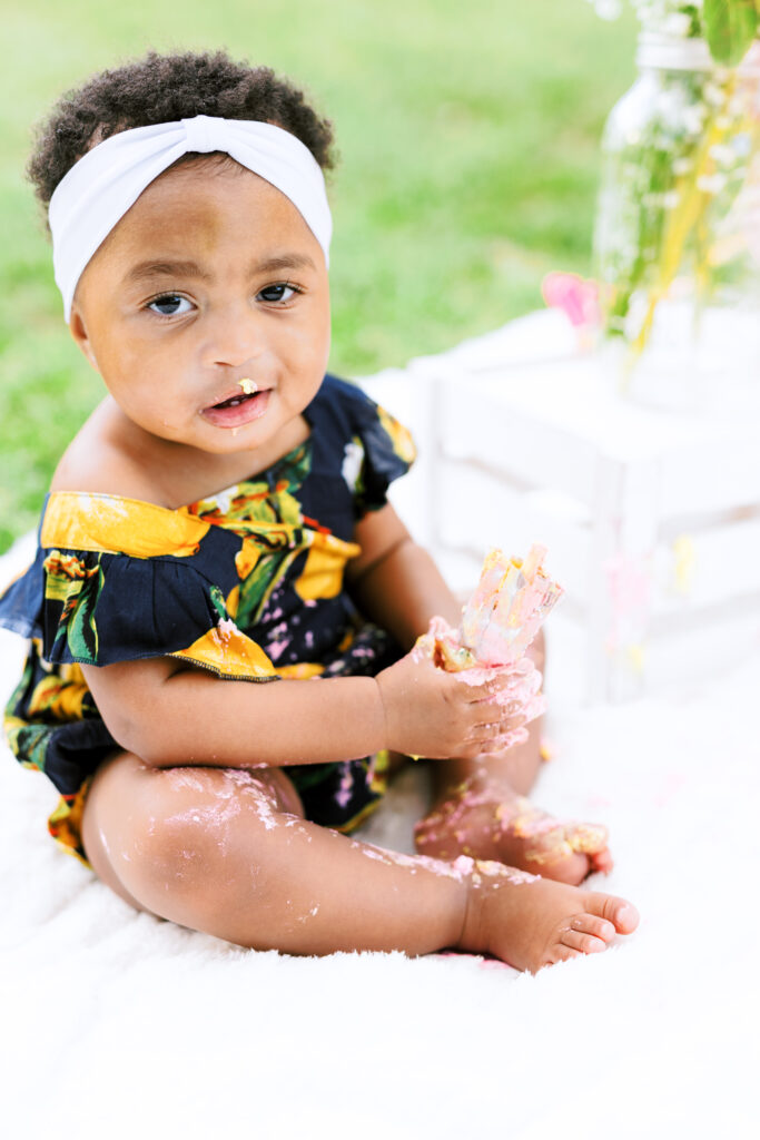 Baby girl licks icing of first birthday smash cake during milestones photo session at Piedmont Park in Atlanta, Georgia